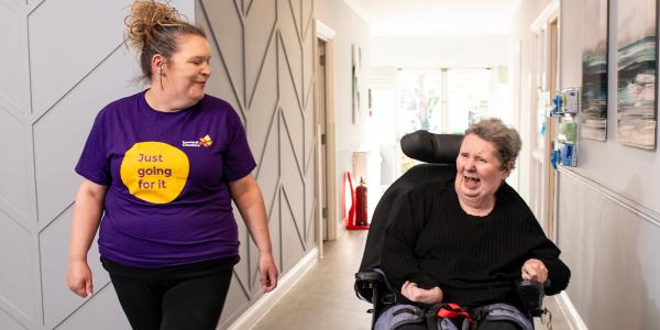 Two Caucasian women going down a corridor one in a wheelchair smiing and the other walking alongside smiling wearing a leonard cheshire branded t shirt