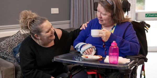 A Caucasian care worker kneeling down next to another female resident smiling holding a cup in her hand