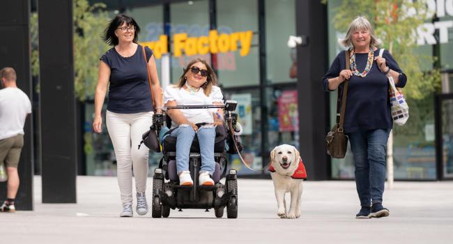 Female wheelchair user with her guide dog and her carer outside some shops