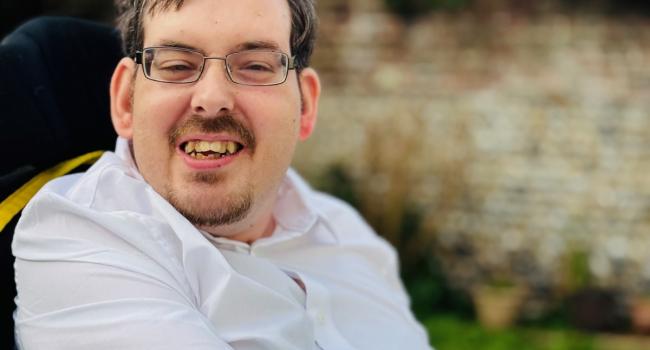 Ashley a Caucasian male with glasses and dark hair and a beard wearing a white shirt sitting in his wheelchair in an outside setting