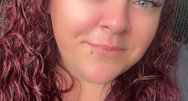 A portrait shot of Tina with reddish brown curly long hair smiling at the camera wearing a grey top 
