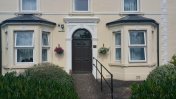 A cream coloured house with arched doorway and a black front door with a slope and handrails in front of it 