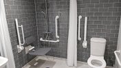 A grey tiled assisted bathroom with shower seat and hand rails along with a toilet 