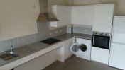 A kitchen space with cream walls and grey wall tiles along with low lying worktops a chrome extractor fan above hob and white appliances such as fridge and washing machine with the door open