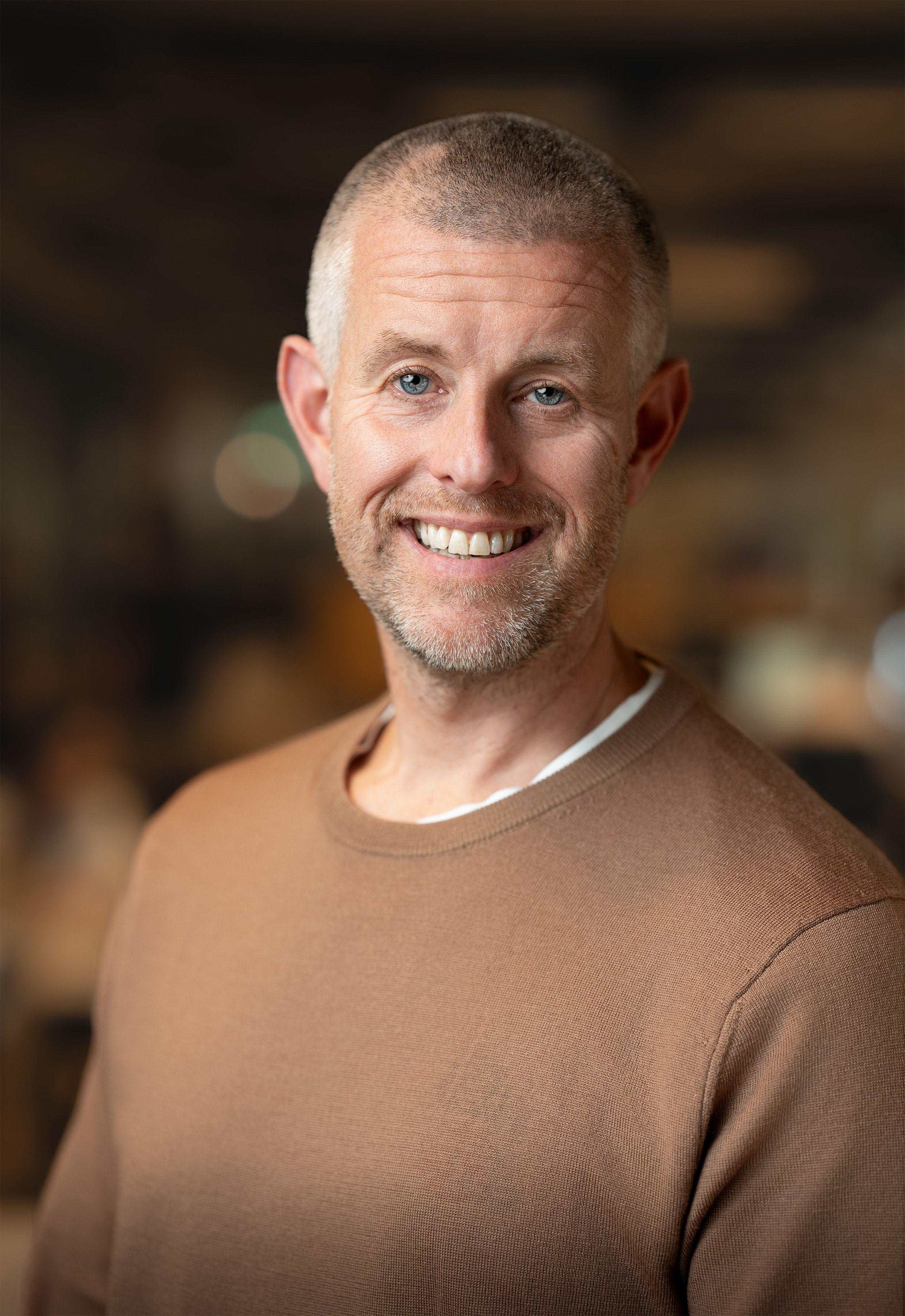 A Caucasian male with grey hair and grey stubble wearing a brown jumper and smiling at the camera