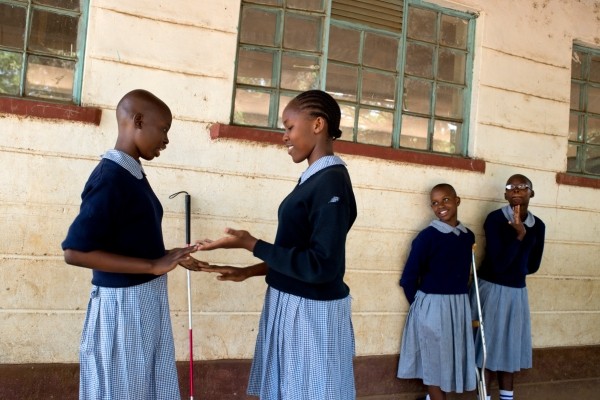 Four girls standing outside a school. Two are facing each other playing a clapping game