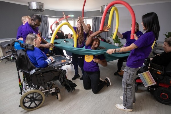 A group of disabled residents in a living area playing with the care workers with foam tubing who are wearing branded t shirts