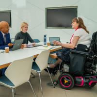 A Caucasian women in a wheelchair sitting opposite a man and a woman around a while modern looking table and chairs in an open plan office space