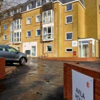 A four storey yellow bricked building and a double doored entrance in front of a forecourt with the Leonard Cheshire branded sign as you drive in reading 'Avila House'