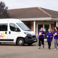 Three female care workers walking through the forecourt in front of a Leonard Cheshire branded minibus 