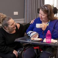 A Caucasian care worker kneeling down next to another female resident smiling holding a cup in her hand