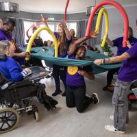 A group of disabled residents in a living area playing with the care workers with foam tubing who are wearing branded t shirts