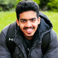 An asian male with black hair and a moustache wearing all black and sat on garden bench with his hands together smiling at the camera with a garden lawn behind him
