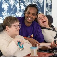 Two caucasian female residents in wheelchairs either side of a female african care worker wearing a purple Leonard Cheshire branded t shirt smiling 