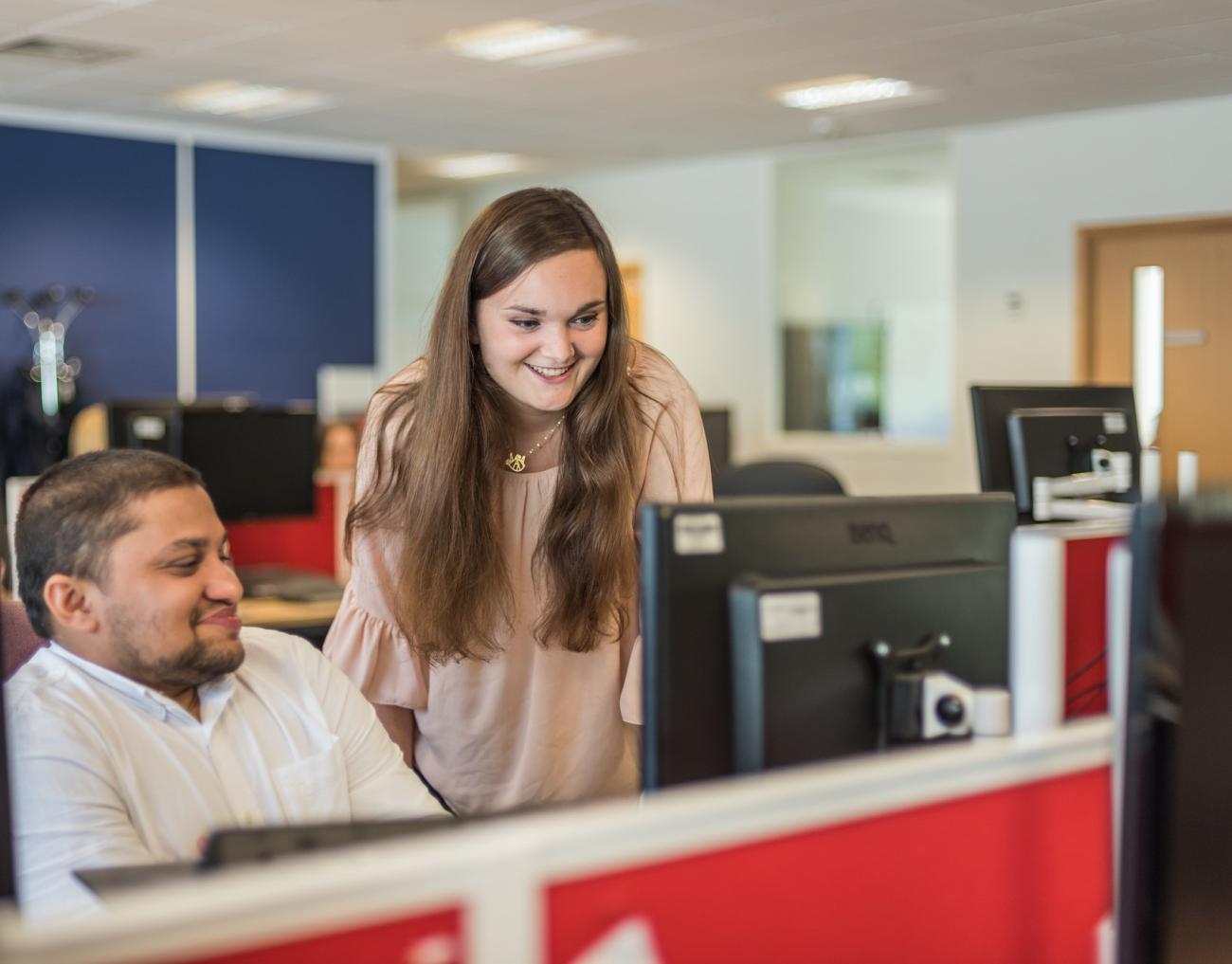 A man sitting at a desk with a woman standing next to him in an office, they're both looking at a screen