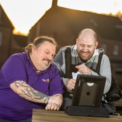 Two men laugh while watching something on a tablet